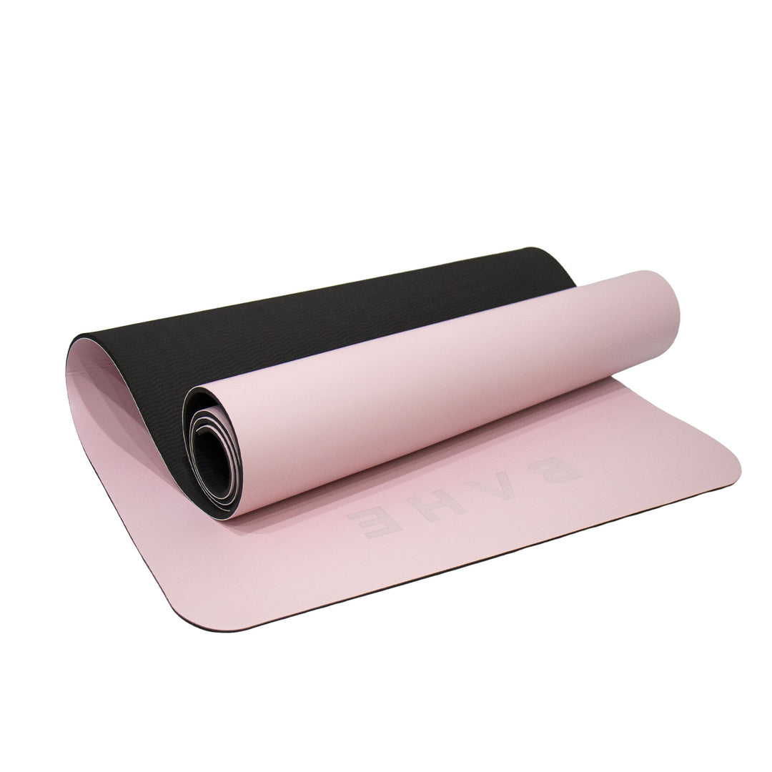 A pink yoga mat with a black textured underside, shown partially unrolled on a clean white background. The brand name &