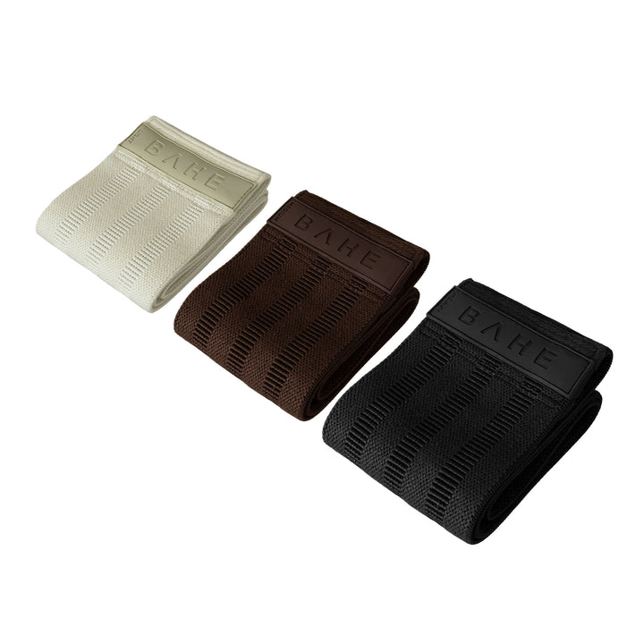 Three booty band resistance straps for pilates in cream, brown, and black, arranged diagonally on a solid white background. Each band features a ribbed texture and a small end tab with the brand name &
