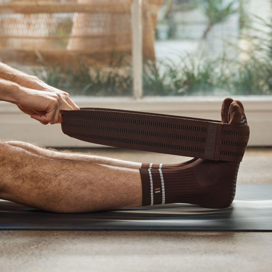 A person stretching a with a dark brown Bahe resistance band in medium resistance, wearing a matching dark brown ribbed sock in a bright indoor setting with a window in the background.