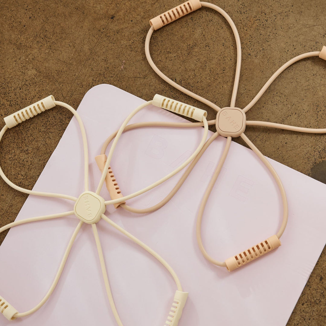 Two beige elastic wrist/ankle exercise bands with textured grip ends, laid on a white sheet over a brown textured surface. The Bahe flow reformer uses resistance training for a pilates workout. 