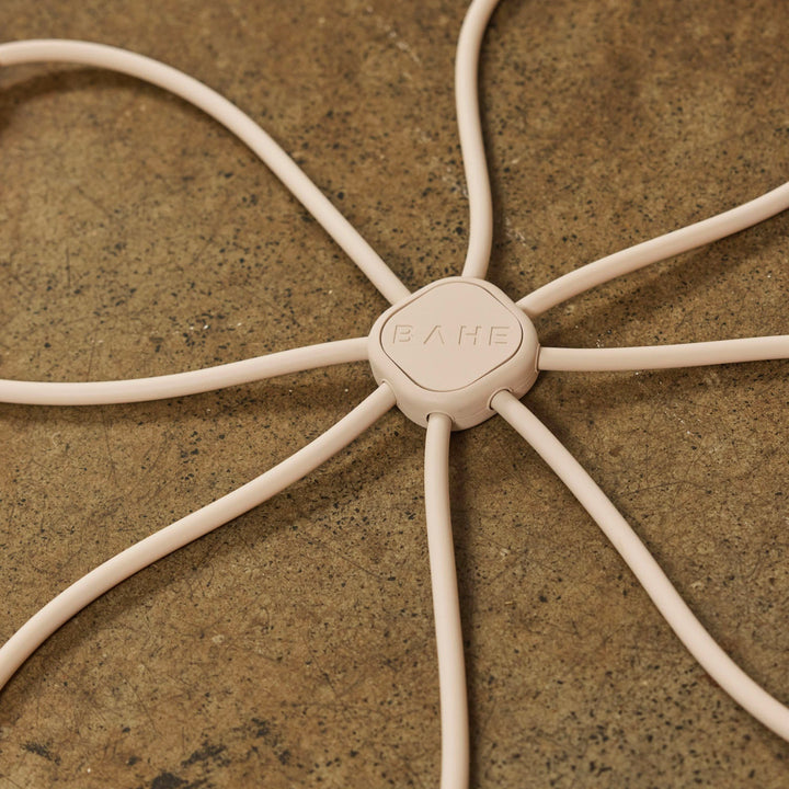 A close-up of a light beige flow refomer by Bahe which mimics the movement of a reformer carriage, showing thin strands radiating outward, laid on a textured brown stone surface.